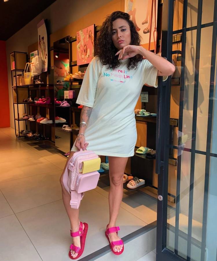 Uma mulher branca de cabelos enrolados posa na porta de uma loja de sapatos ela usa um vestido estilo camiseta branca e uma papete rosa, ela segura uma bolsa rosa