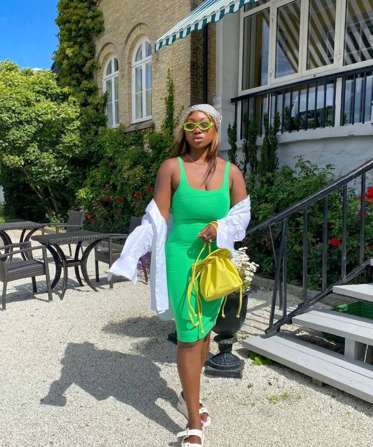 uma mulher negra posa em frente a uma casa. Ela está usando um vestido verde, uma camisa branca caida no ombro um lenço na cabeça 