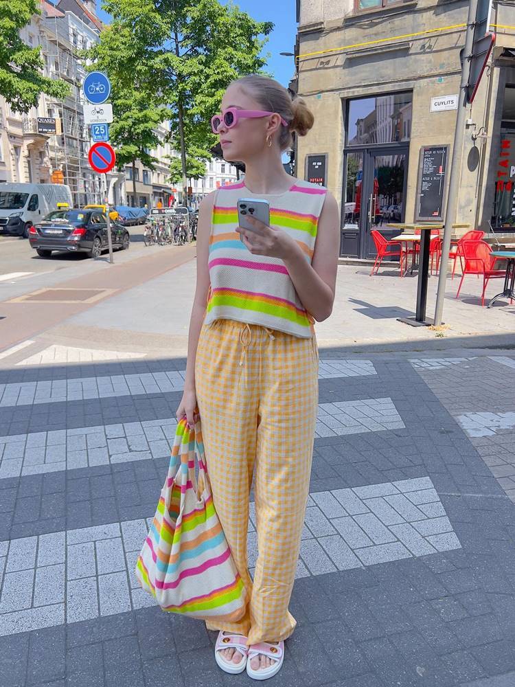Mulher de pele clara em rua usando look colorido, composto por regata listrada neon, calça quadriculada amarela, bolsa listrada neon, óculos de sol pink, e sandálias papete, uma das tendências do verão 2024