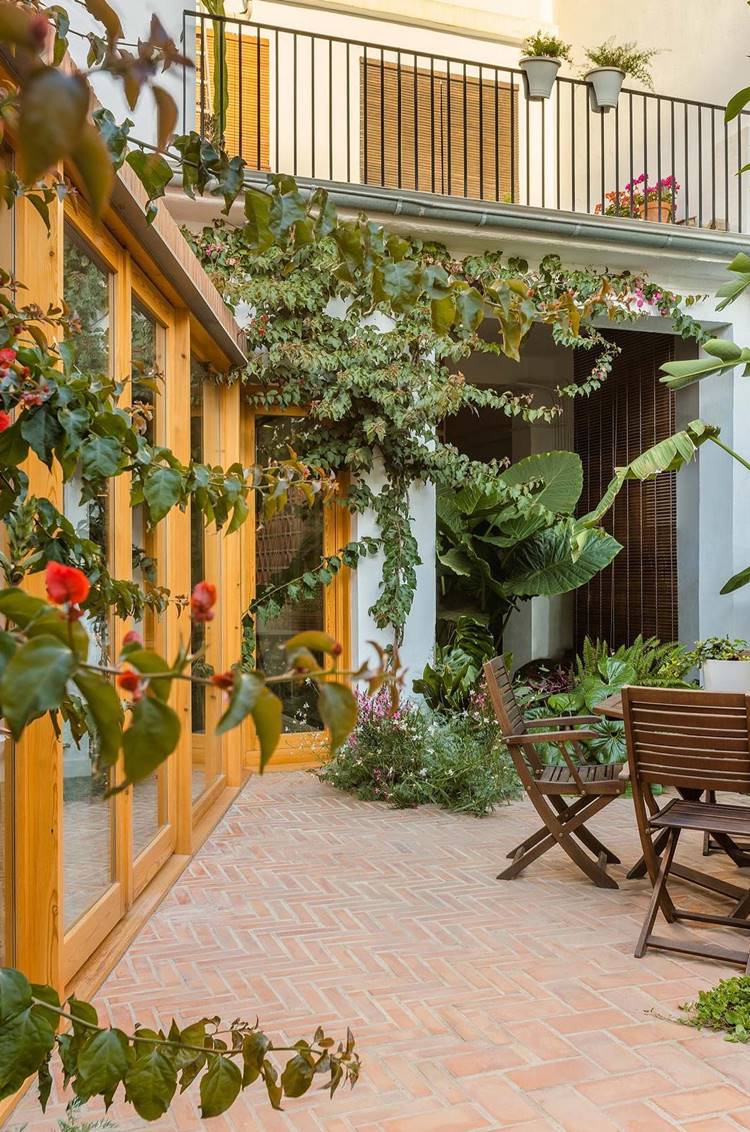 Imagem de fachada de casa simples com muitas plantas decorando, trepadeiras, de folhas grandes, em vasos e pequenas. Mesa de madeira