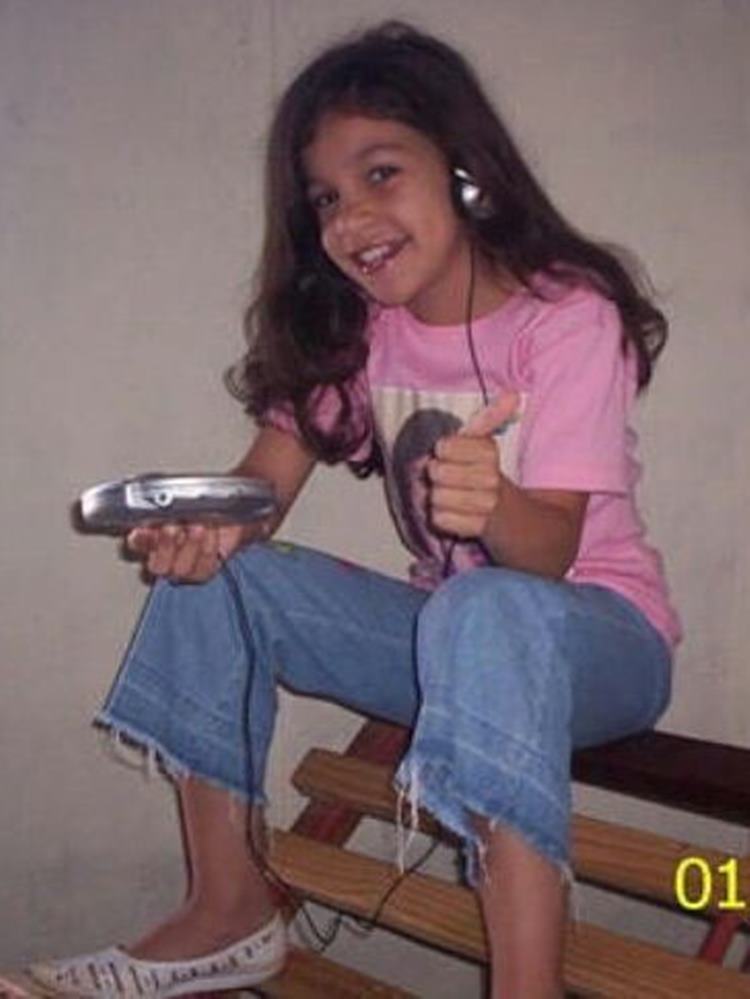 Foto de Priscilla Alcantara quando criança, sentada em cadeira de madeira, usando camiseta rosa com sua foto, capri jeans, sapatilha e usando cd player portátil