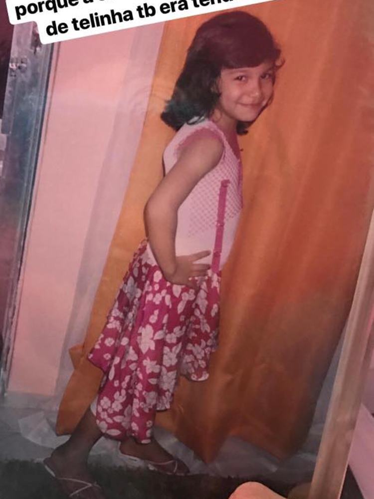 Priscilla Alcantara quando criança, usando blusa rosa com telinha, saia midi rosa florida, sapatilha, de frente a cortina de tecido