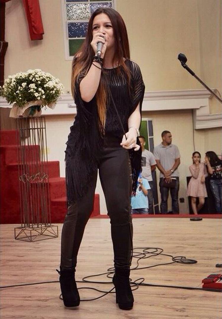 Priscilla Alcantara jovem, com look preto, cantando em igreja 
