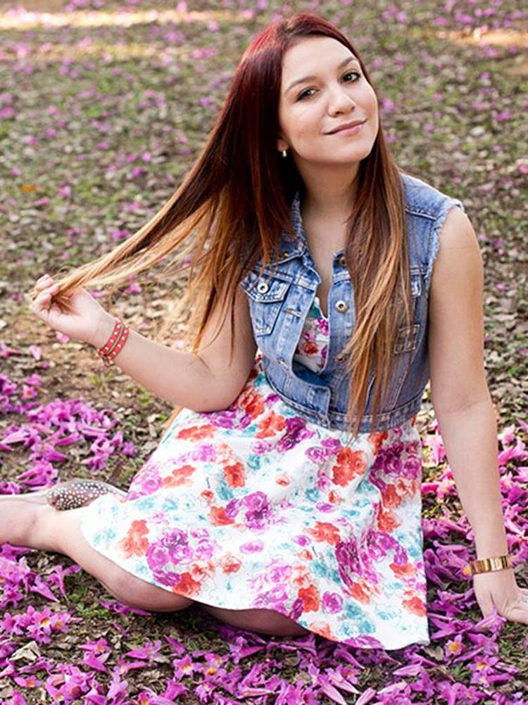Priscilla Alcantara jovem, sentada em gramado, usando vestido florido e colete jeans
