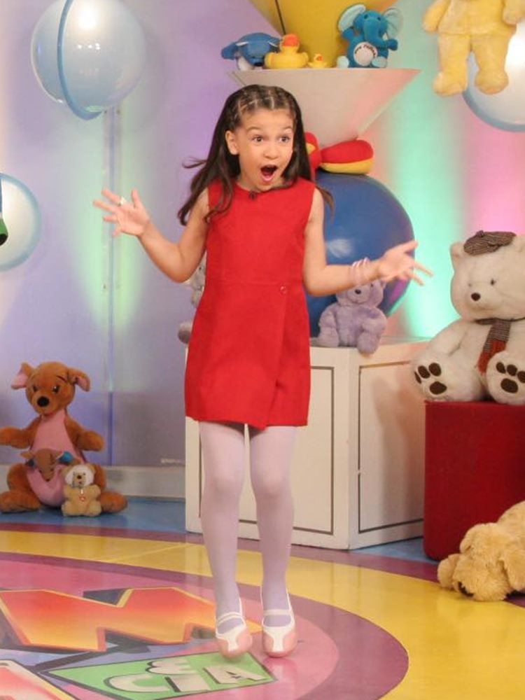 Priscilla Alcantara quando criança apresentando o Bom Dia e Companhia, usando vestido vermelho, meia calça e sapatilha, em cenário com ursos de pelúcia