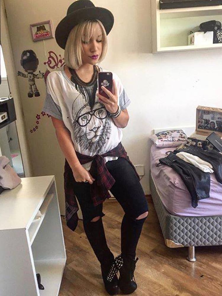 Priscilla Alcantara jovem, com cabelo platinado médio, em selfie, usando camiseta com estampa de leão, calça preta, salto alto, em quarto