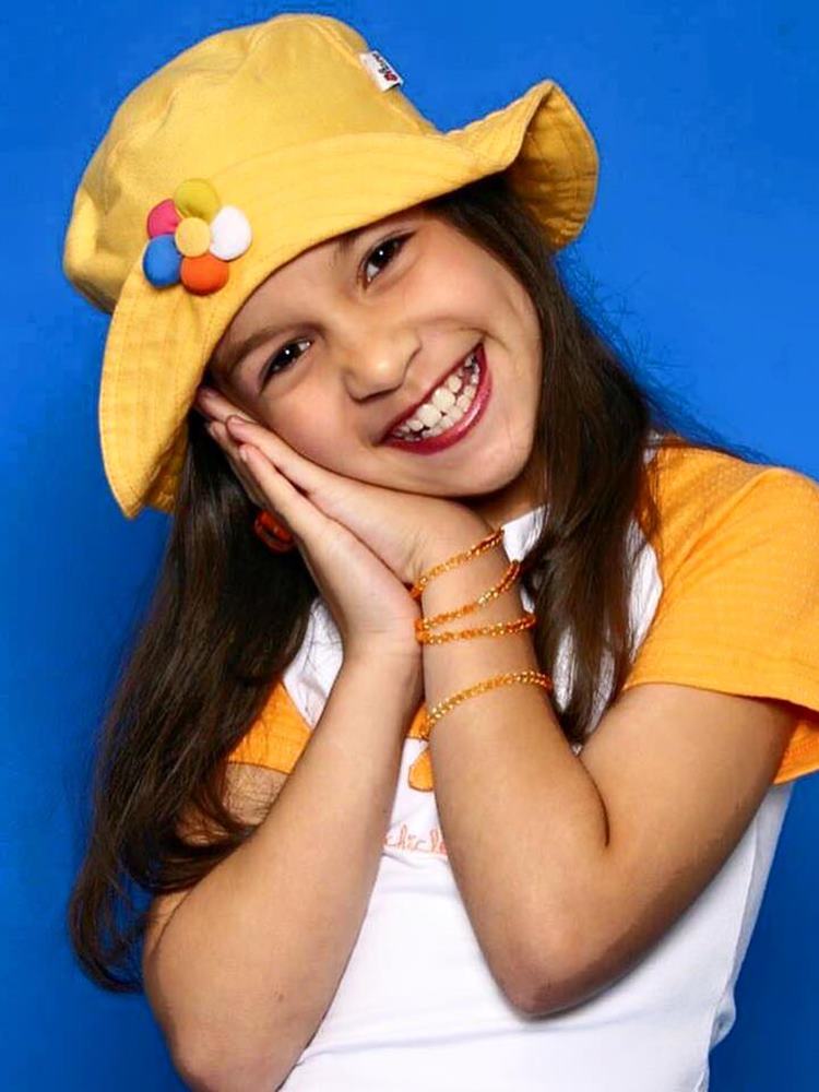 Priscilla Alcantara quando criança, usando roupa branca e amarela, chapéu amarelo com flor colorida e pulseiras de miçangas