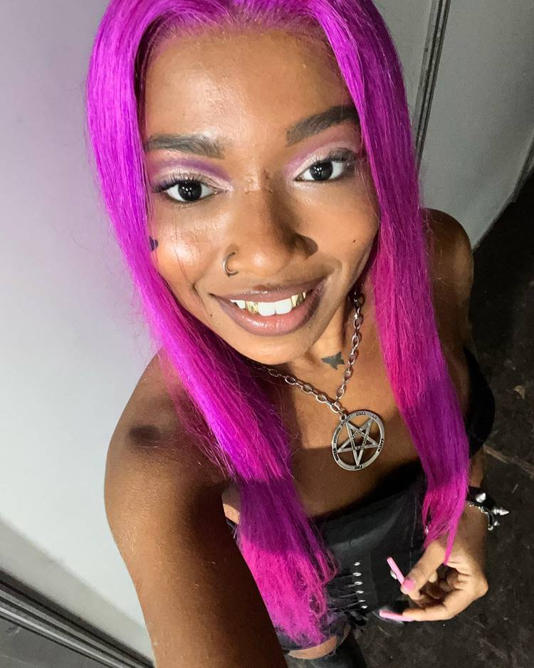 Selfie da affair de MC Cabelinho, cabelo pink, com make esfumada rosa, sorrindo exibindo ouro nos dentes, piercing no nariz, usando corrente com estrela de davi no pescoço. Ela veste roupa preta ousada