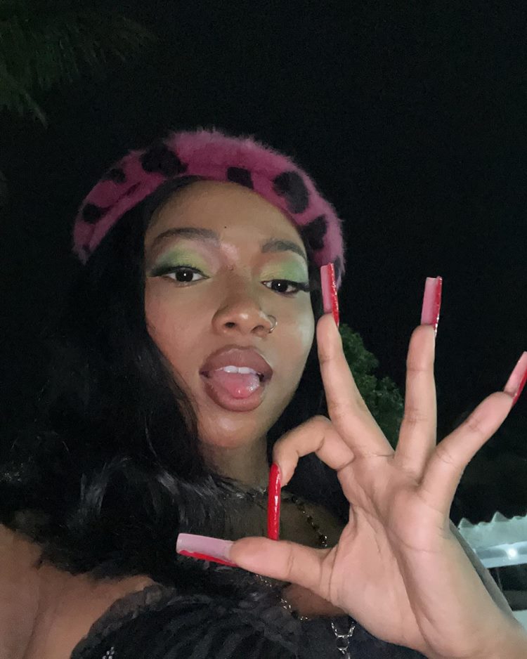 Selfie de Slipmami, fazendo sinal de círculo com os dedos indicador e polegar. Unhas bem grandes vermelhas. Ela usa lace preta até o ombro. Make verde e boia de pelinho