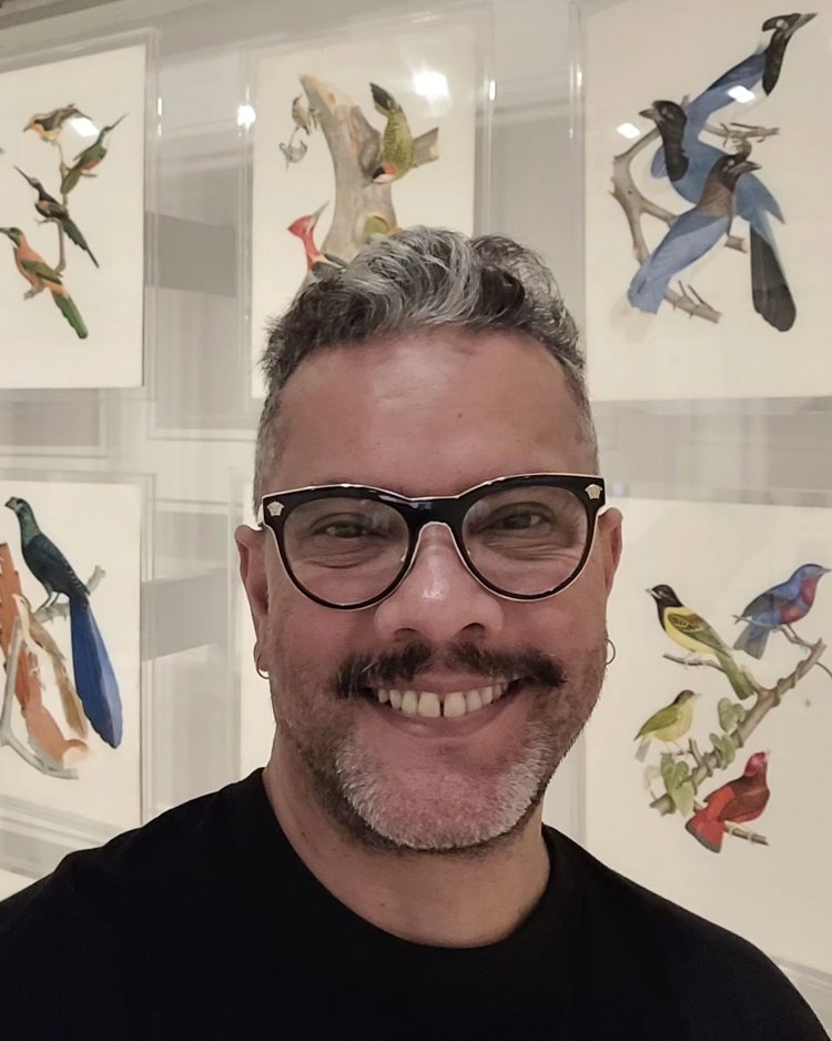 Estilista Ronaldo Silvestre, criador de alguns looks do desfile da Sou de Algodão no SPFW N56, de óculos e camiseta preta, em frente a pinturas de pássaros