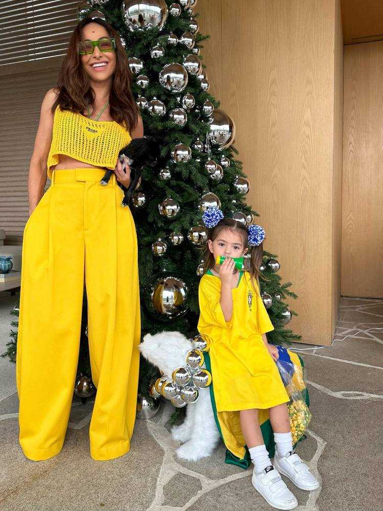Sabrina Sato de cropped e calça amarela, com Zoe de camisa amarela da Seleção Brasileira em frente à arvore de Natal. 
