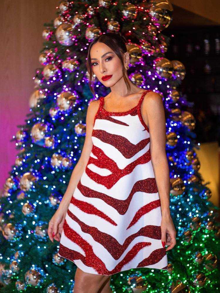 Sabrina Sato usando vestido curto de alças branco com detalhes de zebra em vermelho, em frente á arvore de Natal. 