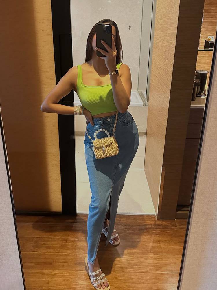 Mulher de pele média e, foto no espelho de banheiro, usando top verde + saia longa jeans, + sandália com pérolas + bolsa de palha com pérolas