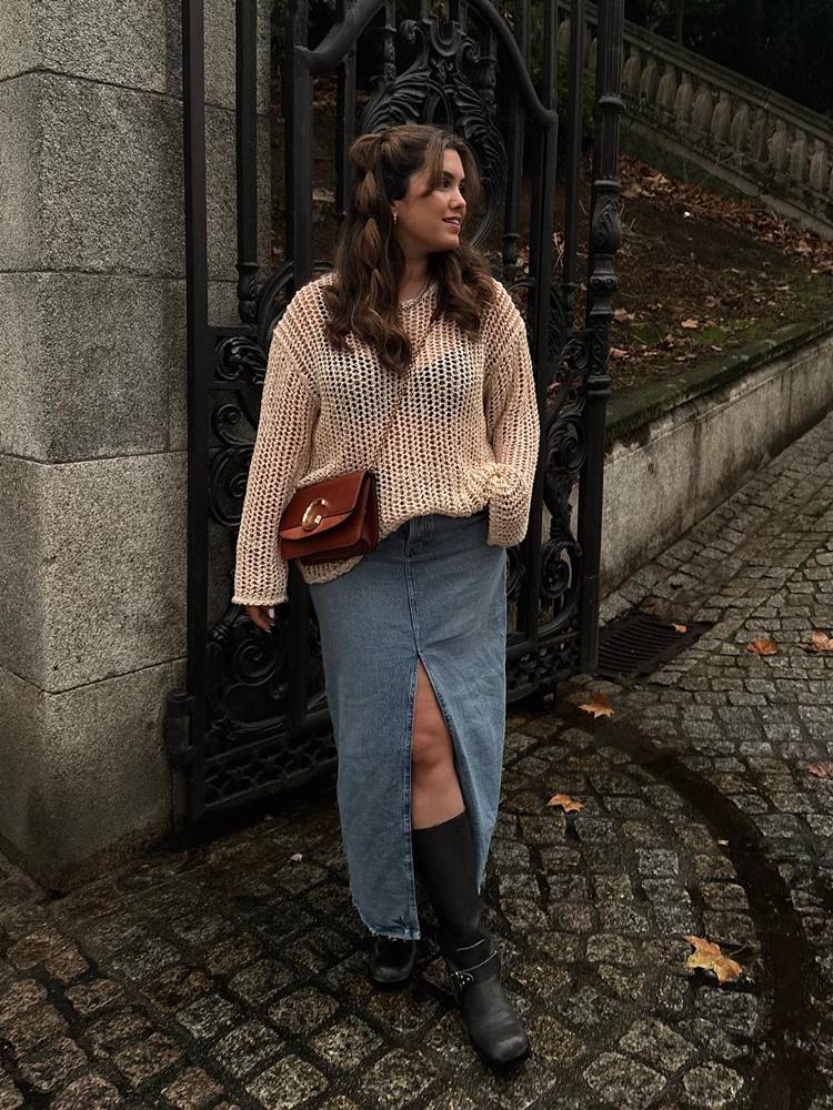 Mulher de pele média usando saia longa jeans, tricô bege, bolsa marrom, bota preta em portão