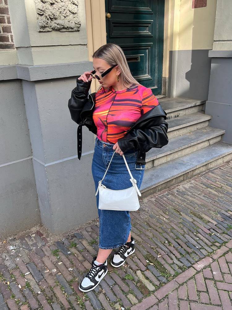 Mulher de pele clara usando body colorido, saia jeans longa, jaqueta de couro preta, bolsa branca e tênis nike em passeio de rua
