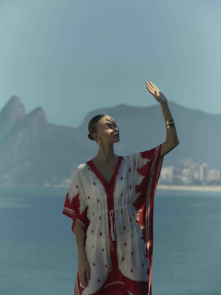 Modelo Yana Trufanova usando uma saída de praia branca e vermelha da coleção Cruise Collection 24 da Lenny Niemeyer. A modelo está de cabelo preso e com a mão levantada em direção ao sol. A paisagem é composta pelo mar e morro cariocas. 