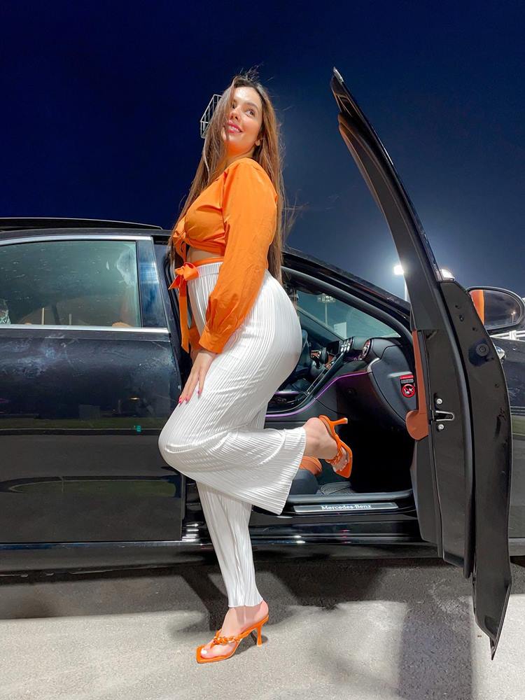 Mulher de pele média em porta aberta de carro, usando cropped laranja, calça branca e sandália flip flop de salto laranja