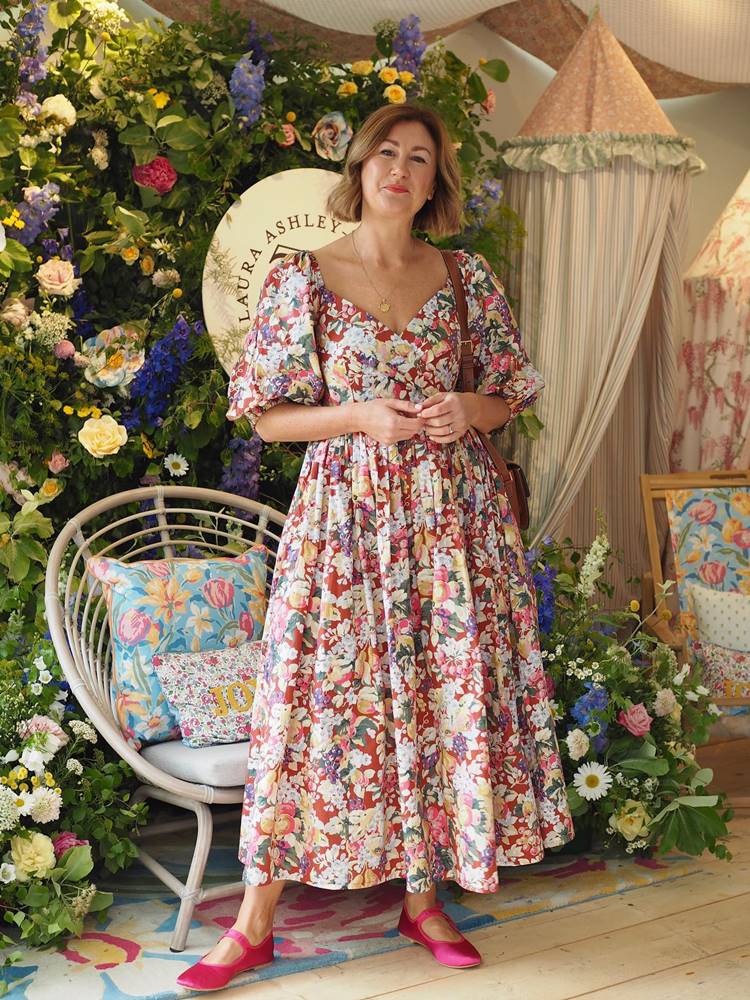 Mulher de pele média em cenário com cadeira e flores de loja, usando vestido floral colorido, bolsa marrom e nos pés sapatilhas de balé pink, uma das tendências em sapatos para o verão 2024