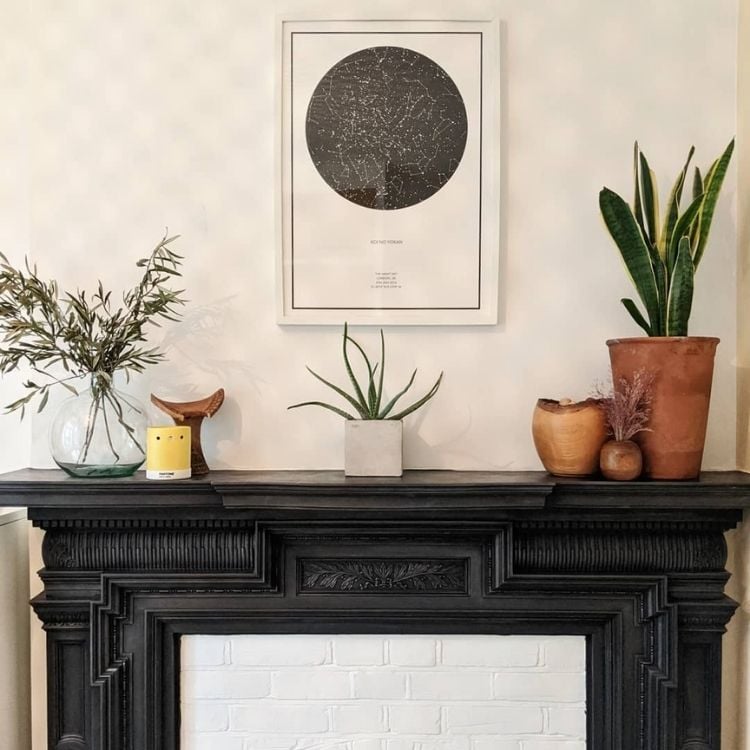 uma lareira preta, com vasos de planta como decoração. Na parede um quadro branco com uma constelação em preto