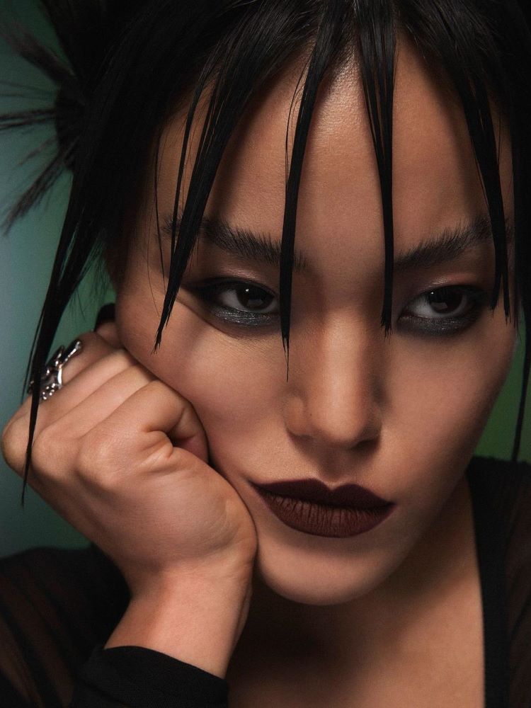 Mulher oriental com cabelo cheio de gel, delineador, sombra abrilhante abaixo da linha d'água e batom marrom escuro.