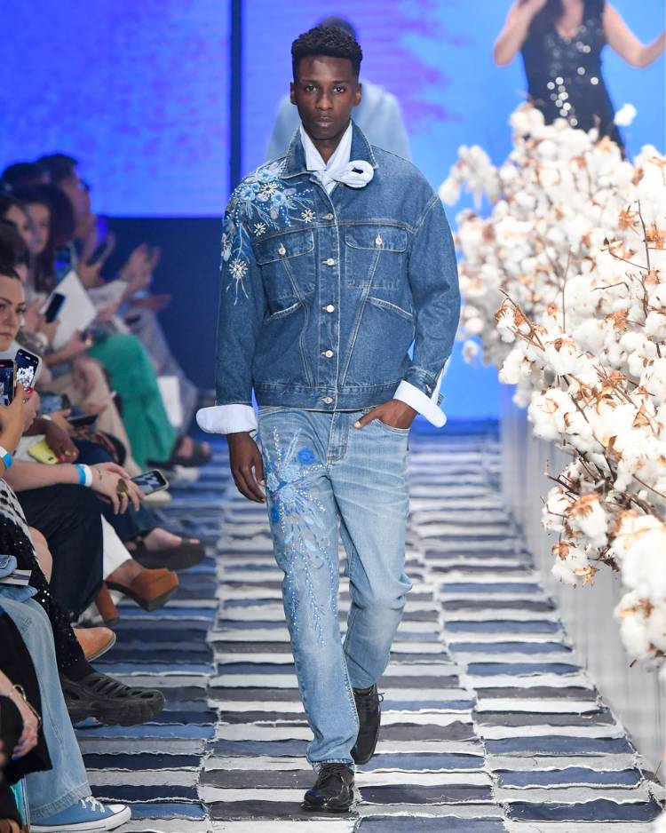 Homem/modelo de pele escura usando look jeans (jaqueta + calça, ambos com bordados da planta algodão) em desfile, passarela com pessoas ao redor e galhos de plantação de algodão decorando
