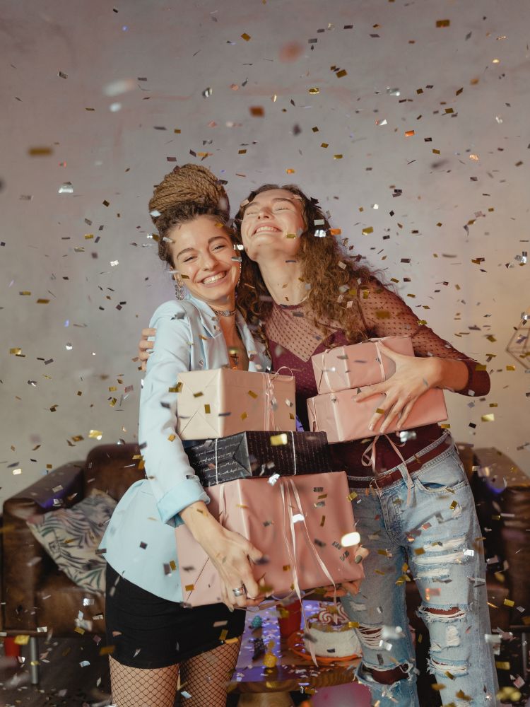 foto de duas amigas segurando caixas de presente em meio a uma chuva de confetes dourados e prateados
