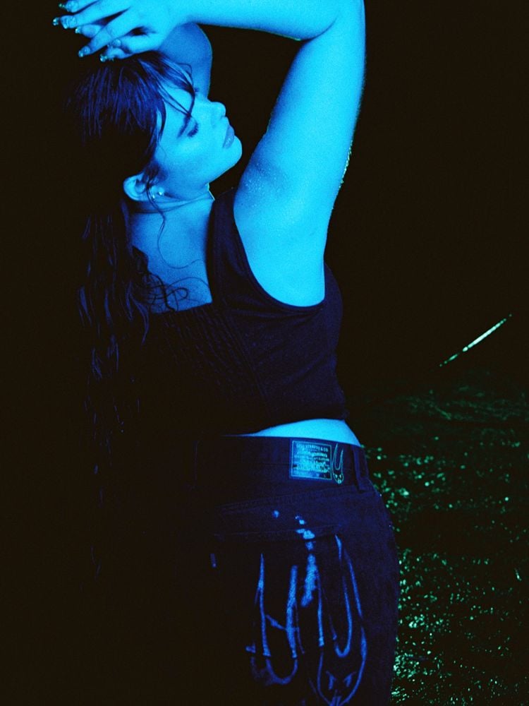 Uma foto com iluminação azul, a atriz posa de costas olhando sobre o ombro. Ela usa uma calça e um corset pretos