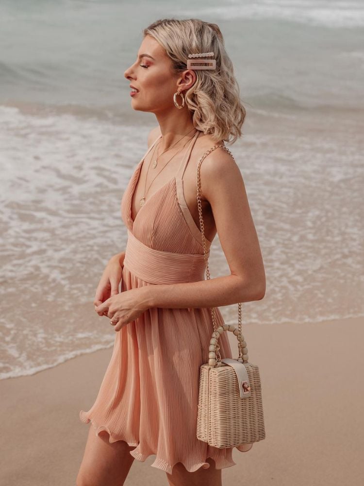 mulher loira na praia usando um vestido pêssego com bolsa de palha