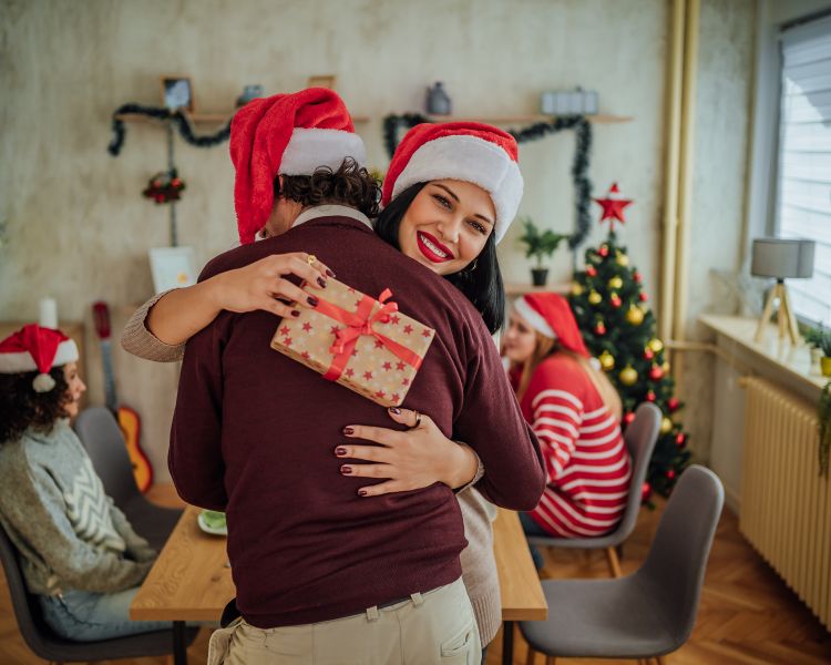 Mulher e homem usando touca de papai noel se abraçando. Ela segura caixa de presentes. Ao fundo, outras duas mulheres com toucas de papai noel sentadas em uma mesa com decoração de Natal ao redor