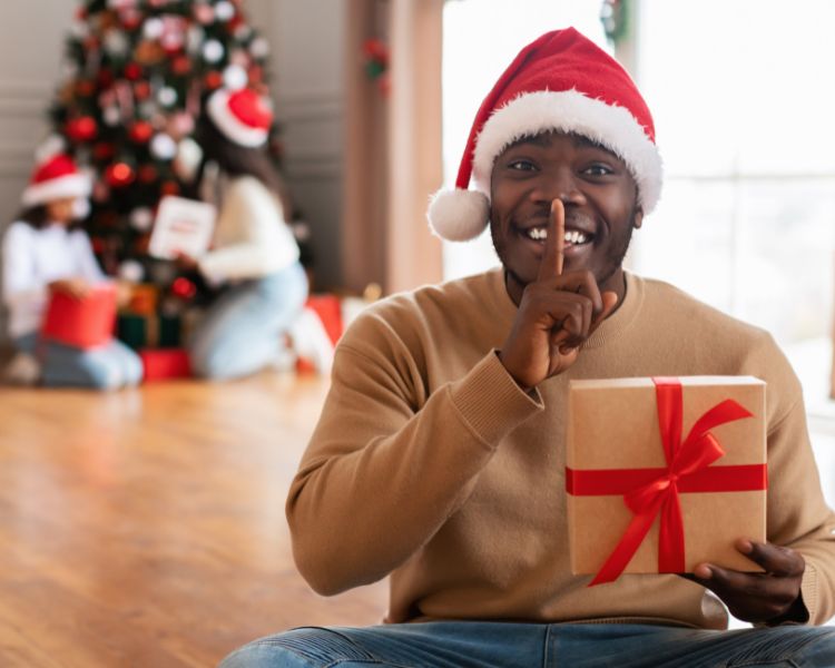 Homem negro fazendo sinal de silêncio com o dedo na boca  usando touca de papai noel e segurando caixa de presentes com laço vermelho. Ao fundo, árvore de Natal e meninas com caixas de presente ao redor