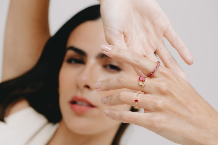 Mulher de pele clara e cabelo escuro usando anéis com pedras vermelhas de Thaís Vilela. 