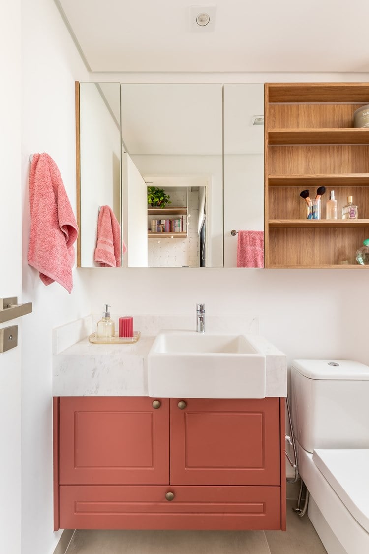 Banheiro do quarto da irmã, com pia rosa, vaso sanitário e armário suspenso com espelho e prateleiras com itens de beleza
