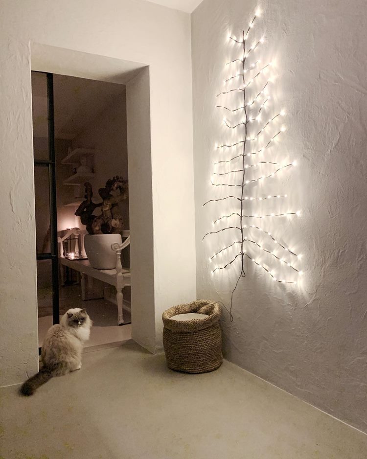 Casa com decoração minimalista com árvore de Natal minimalista na parede, no chão, gato e saco de juta; No cômodo ao lado, banco com vaso de flor e prateleiras