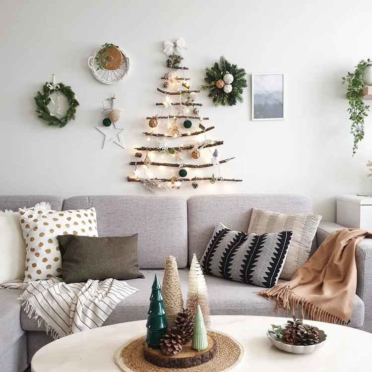 Sala com sofá cinza com almofadas e mantas, mesa de centro decorada com árvores de Natal. Na parde de fundo, quadros, guirlandas e árvore-de-natal de parede feita de galhos