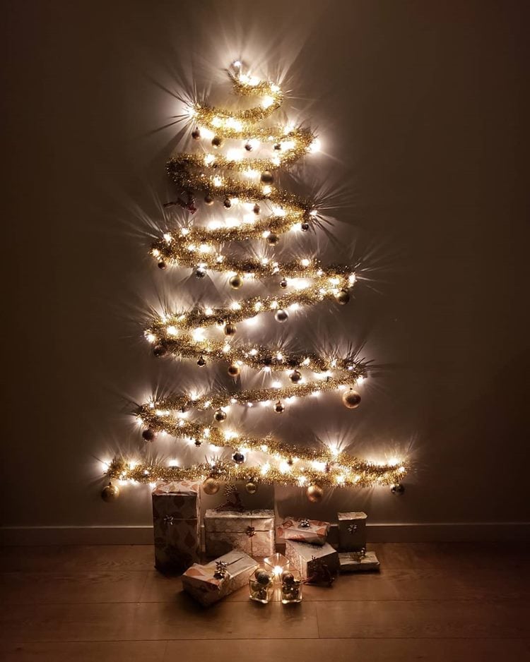 Árvore-de-natal simples feita de festão dourado, bolas natalinas e pisca-pisca na parede, com caixas de presentes abaixo, no chão