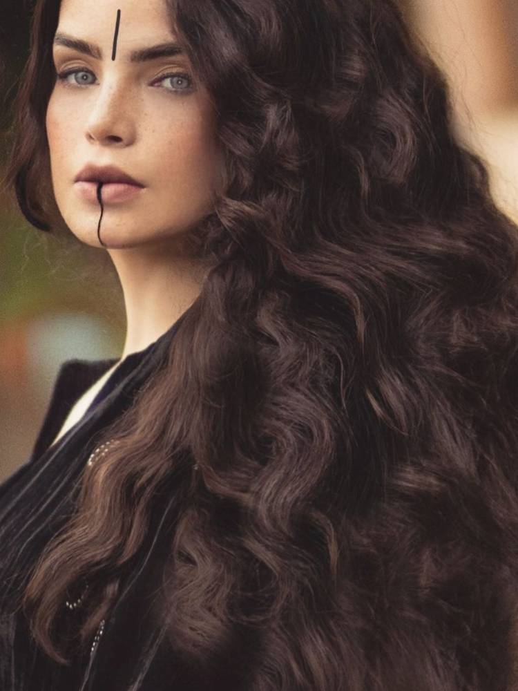 Mulher de pele clara e cabelos escuros mística, com linha reta preta no rosto e roupas pretas.