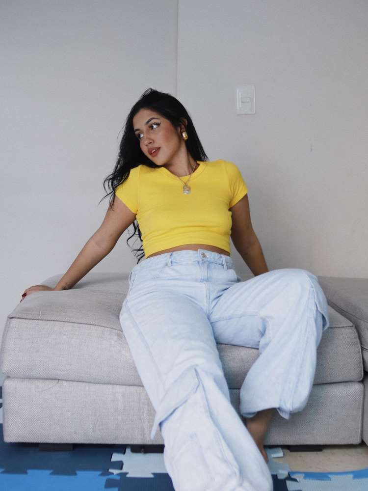 Mulher de pele clara e cabelo escuro sentada em uma sofá cinza e usando calça jeans larga e baby look amarela. 