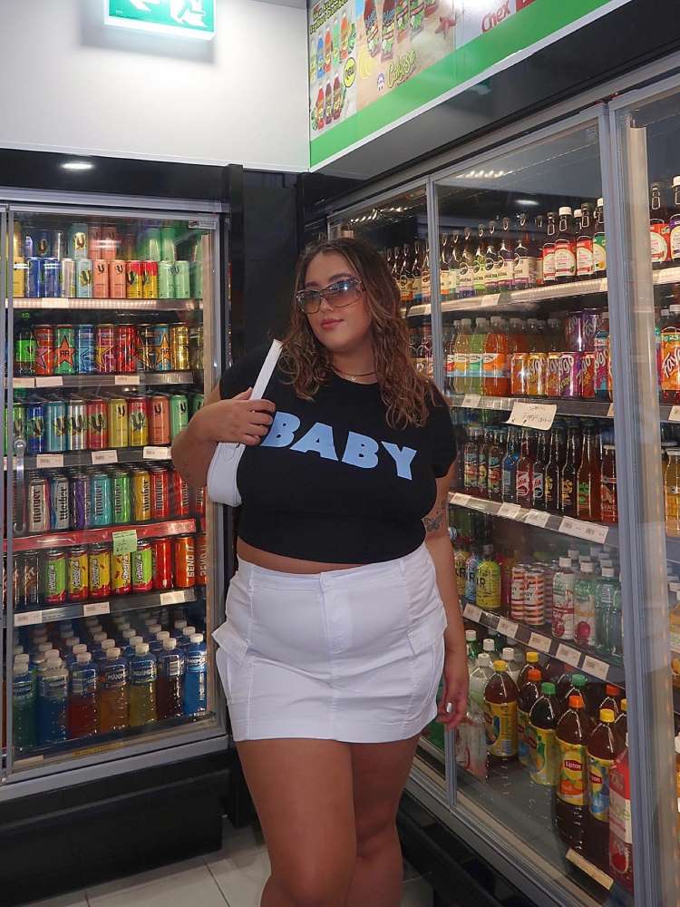 Mulher de pele morena no supermercado usando baby look preta escrito "baby" em azul e saia branca. 