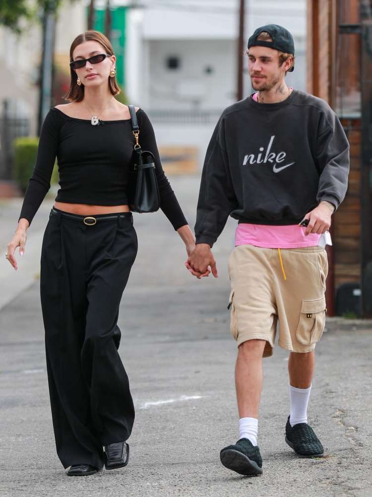 Hailey Bieber usando calça preta e blusa baby look preta, andando na rua acompanhada de Justin Bieber, que usa moletom preto e bermuda bege. 