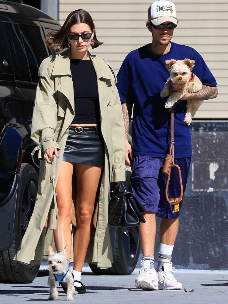 Hailey Bieber usando camiseta baby look preta, minissaia azul escura, casaco cinza, acompanhada e Justin Bieber, que segura um cachorro.