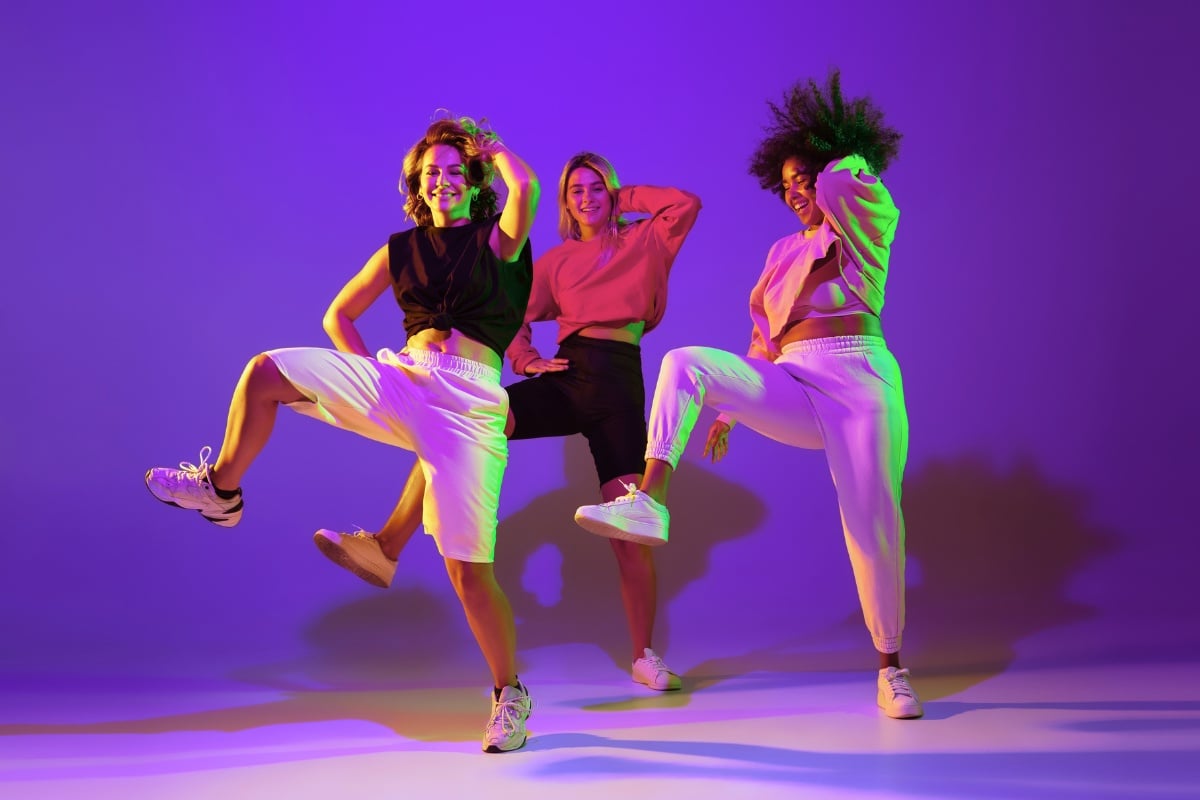 Foto de 3 mulheres fazendo passo de dança em fundo roxo com detalhes neon. Elas usam roupa esportiva