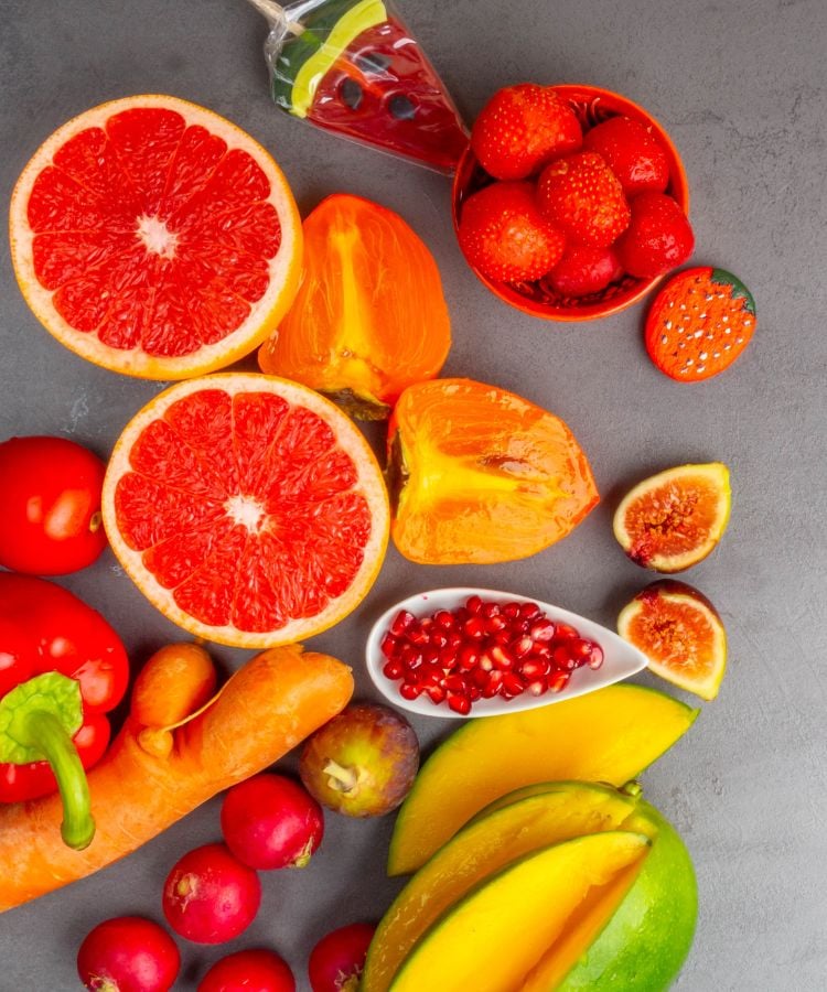 A imagem apresenta uma variedade de frutas e vegetais frescos e coloridos. Duas metades de grapefruit vermelha estão visíveis. Pedacinhos de caqui laranja brilhante estão próximos às grapefruits .Uma tigela pequena está cheia de morangos frescos e suculentos.Pimentões vermelhos inteiros adicionam outra camada de cor à composição. Uma cenoura laranja está presente na imagem. Grãos de romã são mostrados em um pote branco ovalado.