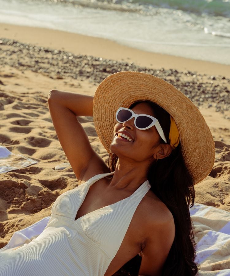 Uma mulher está relaxando e fazendo bronzeado saudável em uma praia ensolarada. Ela está usando um vestido branco e um chapéu de palha grande
