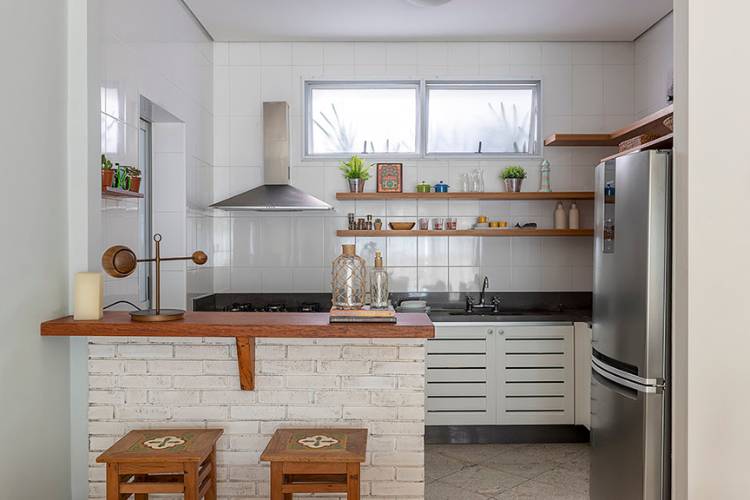 Cozinha com balcão americano, tijolinho à vista, prateleiras e banco em madeira, armários brancos, geladeira, coifa e itens de decoração. 
