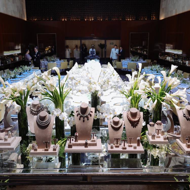 Decoração do almoço da joalheria Cris Porto com joias e jarros de vidro com orquídeas e copos de leite brancos, ao fundo, as mesas-postas dos convidados e garçons