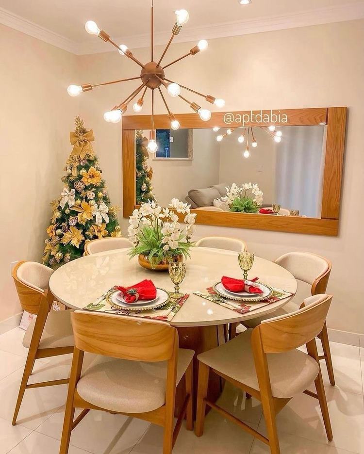Uma mesa redonda com 6 cadeiras, mesa-posta com 2 pratos, arrajo de orquídea. No canto da parede, uma árvore de Natal com enfeites dourados. Na parede, um espelho grande retangular. Acima, lustre com várias lâmpadas pequenas.