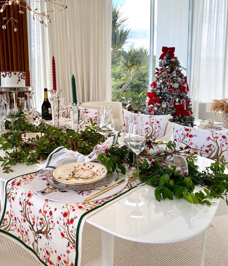 Mesa para ceia de Natal decorada com ramos de planta de verdade. A mesa posta possui toalha e almofadas nas cadeiras com estampa de rena de Natal.