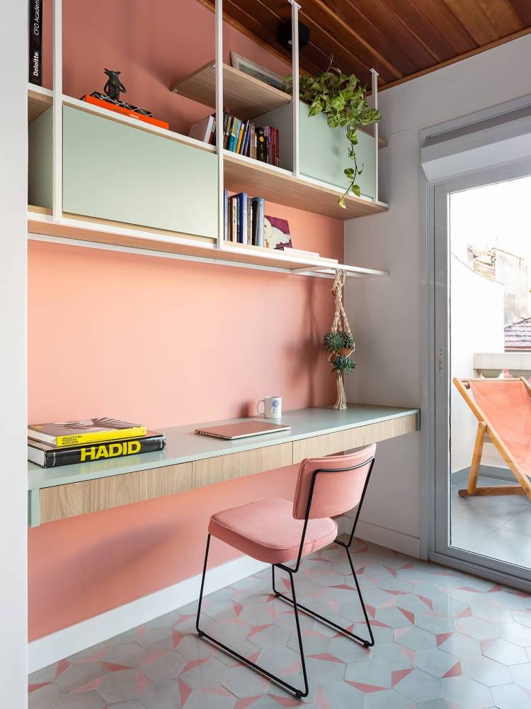 Home office em rosa, cuja estante possui partes abertas e fechadas, com plantas e livros. 