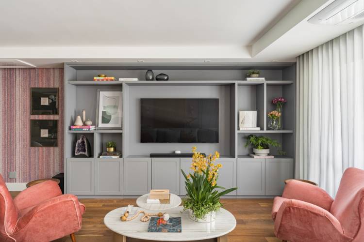 Sala de estar em cinza e rosa, com poltronas rosas, mesa de centro com plantas, estante cinza com TV e diversos itens de decoração, como livros, vasos e quadros. 