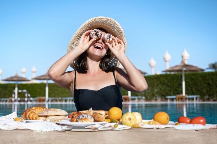 Mulher de pele clara usando biquíni preto, chapéu, colocando donuts sobre os olhos e com uma refeição farta à frente.